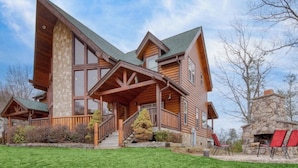 A log cabin with a stone chimney, large windows, and a porch with red chairs. - Luxurious 4BD Lodge with HOT TUB! Fireplace! (Sevierville)