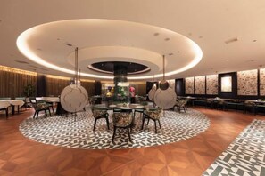 A hotel lobby with a central round table surrounded by chairs, a patterned carpet, and a modern ceiling design.