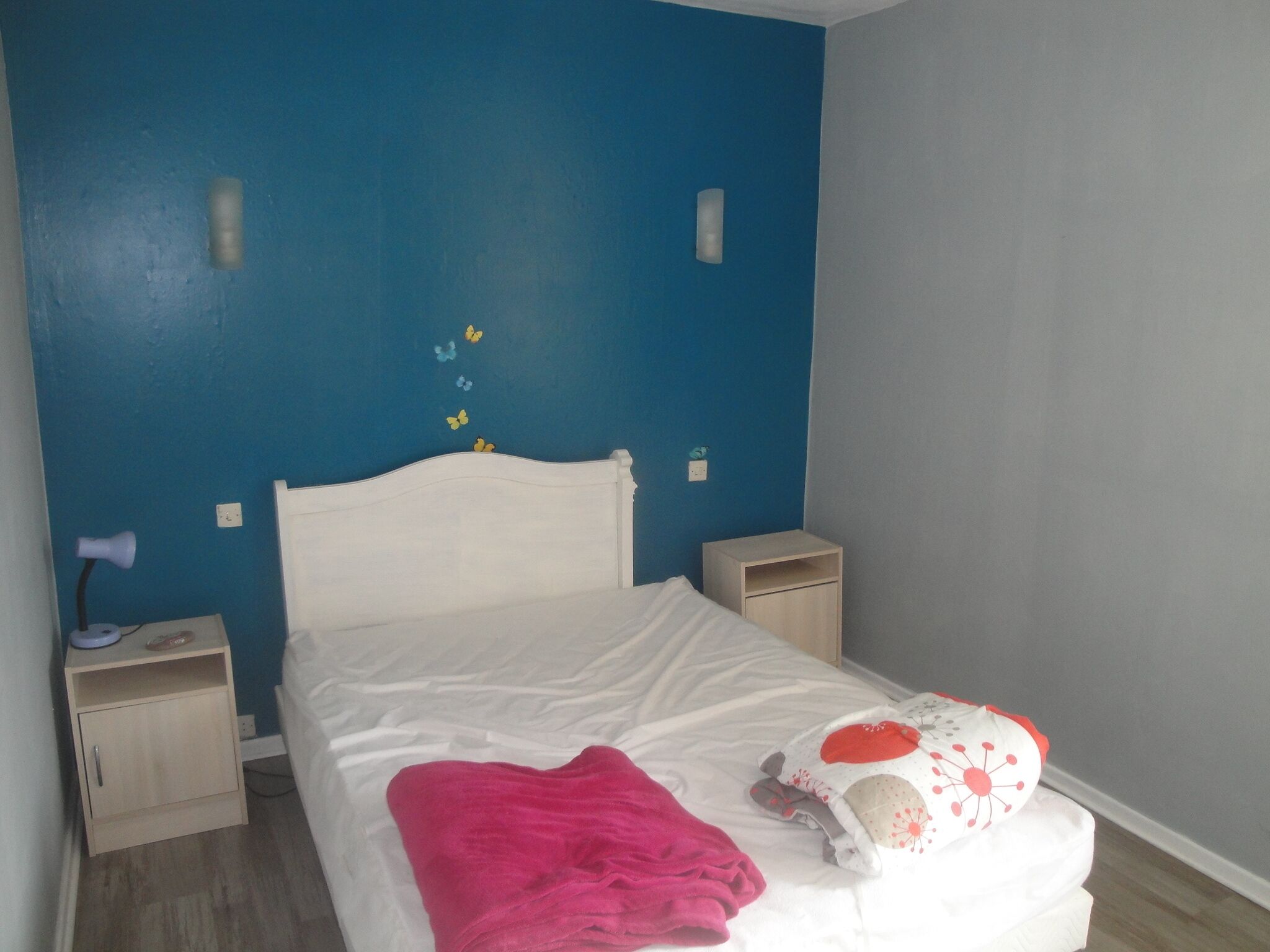 Een slaapkamer met een wit bed, een houten nachtkastje, een bijzettafeltje en een blauwe muur versierd met vlinderstickers.