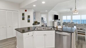 Una cocina moderna con armarios blancos, encimera negra y un lavamanos empotrado. Hay un ventilador de techo y una ventana grande con vistas al océano.