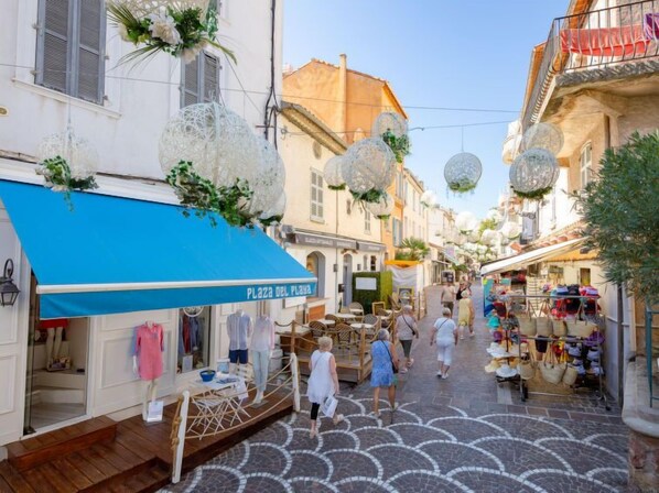 A narrow street with shops and outdoor seating, decorated with hanging spherical ornaments and floral arrangements.