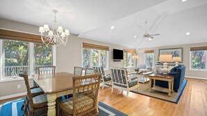 A spacious living room with a wooden dining table, wicker chairs, a blue sofa, a ceiling fan, and large windows with blinds. - Pelican Place - Golf cart included 704 Swordfish (Fripp Island)