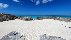 A beach with white sand and dark rocks in the foreground, leading to a clear blue sea under a bright sky. - Bakari House - Luxury Oceanfront Villa w/ Spectacular View, Pool, Spa and Dock (Hartswell)