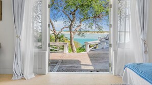 A room with a large glass door leading to a wooden deck overlooking a beach. - Bakari House - Luxury Oceanfront Villa w/ Spectacular View, Pool, Spa and Dock (Hartswell)