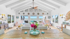 A spacious room with a high ceiling, a wooden dining table, wicker furniture, and a view of the ocean through large windows. - Bakari House - Luxury Oceanfront Villa w/ Spectacular View, Pool, Spa and Dock (Hartswell)