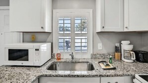 A kitchen with white cabinets, a granite countertop, a stainless steel sink, a white microwave, a coffee maker, and a window with a view of trees. - Snowbirds save 50% Clean , pets, beachside apt. Downtown Flagler Beach -Sleeps 4 (Flagler Beach)