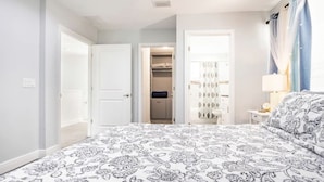 A bedroom with a patterned bedspread, a closet with hanging clothes, and a bedside table with a lamp.