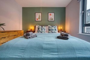 Ein Schlafzimmer mit einem Bett, Kissen, einem Nachttisch, einer Lampe und einem Fenster.