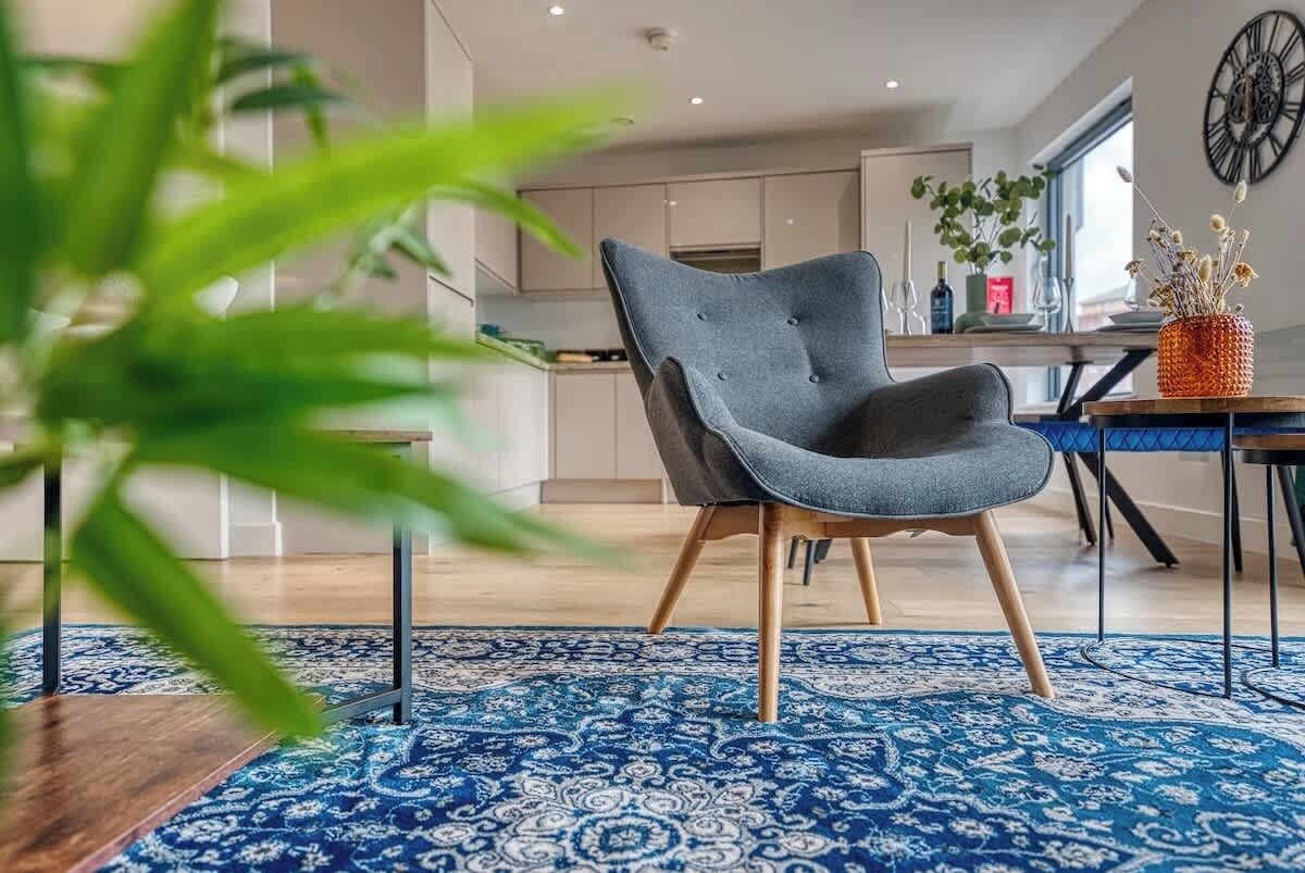 A modern living room with a blue patterned rug, a grey armchair, and a dining area with a blue table.
