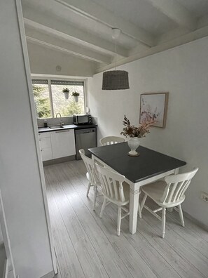 A kitchen with white cabinets, a black countertop, and a window with hanging plants. There is a dining table with white chairs and a vase with flowers.