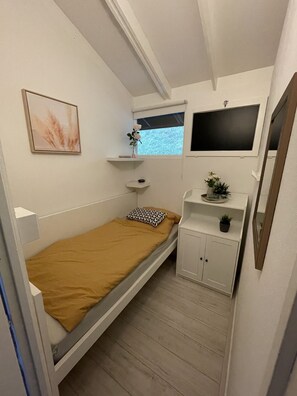 Een kleine, compacte slaapkamer met een eenpersoonsbed, een raam, een wandtelevisie en een klein kastje met een plant.