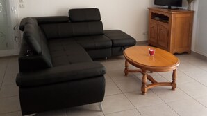 Un salon moderne avec un canapé d’angle noir, une table basse en bois, un téléviseur posé sur un meuble en bois et un climatiseur fixé au mur.