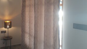 A room with a large window covered by sheer curtains, a wall-mounted lamp, and a small table with a lamp. - Namaste Nilaveli (Nilaveli)