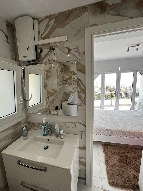 A bathroom with a marble backsplash, a sink with toiletries, a mirror, and a view of a balcony with a white bedspread. - Studio with breathtaking view (Voula)