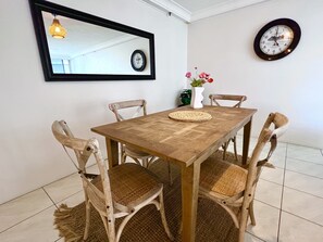 Un comedor con una mesa de madera, sillas tejidas, un jarrón con flores, un reloj de pared y una lámpara colgante.