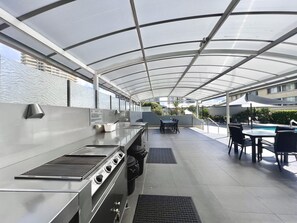 An outdoor kitchen area with a grill, dining table, and chairs, under a covered patio with a view of a swimming pool and buildings. - Ocean View 2BR MainBeach Getaway (Main Beach)