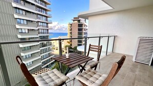 Un balcon avec une table et deux fauteuils confortables, donnant sur un paysage urbain avec des bâtiments et une étendue d’eau.