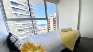 Une chambre d’hôtel avec une grande fenêtre, un lit recouvert de draps blancs, un plaid jaune et une seule fleur jaune posée sur le lit.