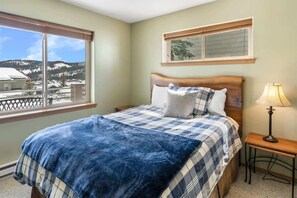 Un dormitorio con un ventanal grande, una cama con una colcha azul, una lámpara de noche y vistas a un paisaje nevado.