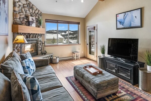 Una sala de estar con pared de piedra, chimenea, ventana grande con vistas, sofá, reposapiés, televisor y aparador.