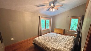 A bedroom with a wooden floor, a bed with a patterned bedspread, a ceiling fan, and two windows with curtains. - Quiet relaxing wooded property. (New London)