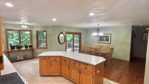 A kitchen with wooden cabinets, a large island, a dining area with chairs, and a clock on the wall. - Quiet relaxing wooded property. (New London)