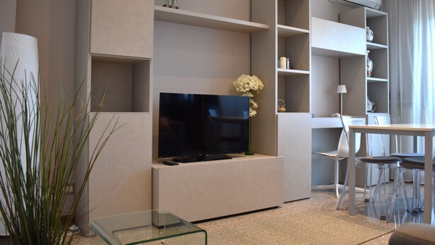 A modern living room with a flat-screen TV, built-in shelves, a dining area with a table and chairs, and a potted plant. - Sundial One. Pradamano (Pradamano)
