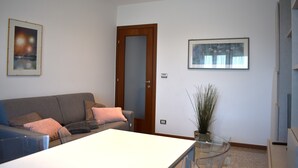 A modern living room with a grey sofa, a white coffee table, and a glass-top dining table. There is a wooden door, a framed picture on the wall, and a potted plant. - Sundial One. Pradamano (Pradamano)