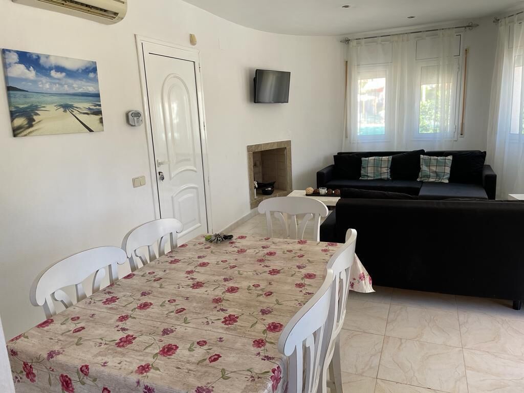 A living room with a black sofa, a white dining table with floral tablecloth, and a TV mounted on the wall.