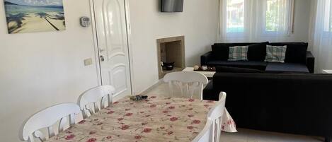 A living room with a black sofa, a white dining table with floral tablecloth, and a TV mounted on the wall.