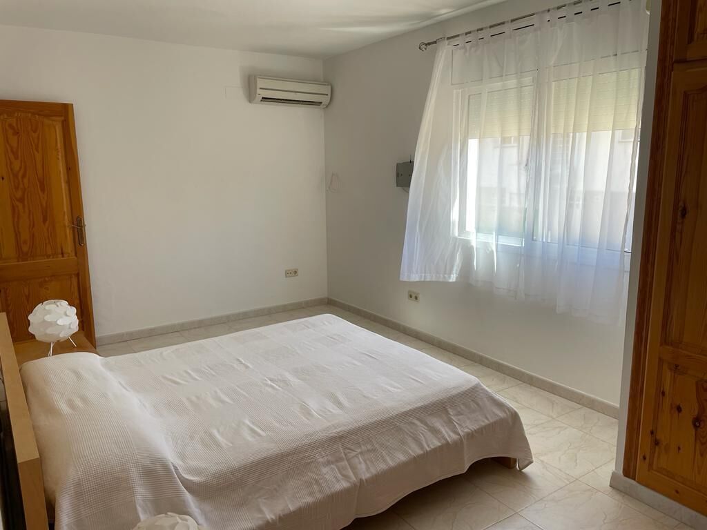 A bedroom with a bed, a wooden wardrobe, a window with sheer curtains, and an air conditioning unit.