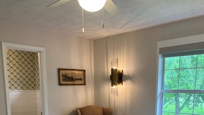 Une chambre avec un grand lit, un ventilateur de plafond, un luminaire fixé au mur, une chaise, un tableau encadré et une fenêtre donnant sur un paysage verdoyant.
