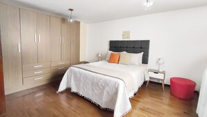 Une chambre à coucher comprenant un lit, une table de chevet, un pouf rouge, une armoire en bois et deux lampes suspendues.