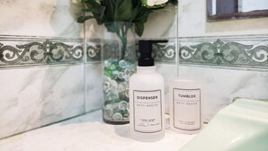 Une salle de bain avec un mur en marbre, un vase rempli de fleurs blanches et deux flacons étiquetés « Essentiels pour le bain » posés sur le comptoir.