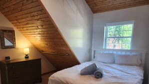 A bedroom with a sloped ceiling, a bed with white linens, a bedside table with a lamp, a dresser, and a window with a view of trees. - The Cottage at Hubbard Lake (Hubbard Lake)