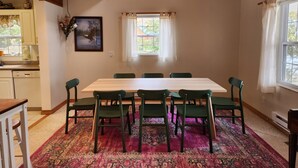 A dining area with a wooden table and six chairs, a window with curtains, and a kitchen visible in the background. - The Cottage at Hubbard Lake (Hubbard Lake)