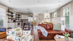 A spacious living area with a brown leather sofa, a patterned armchair, and a coffee table. A kitchen with modern appliances and a dining area with a high chair are visible in the background. - Waterfront Condo, Pool, Walk to Downtown (Saugatuck)