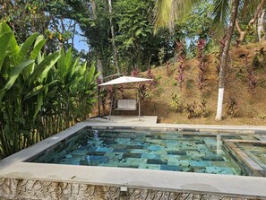 A swimming pool with a tiled deck, surrounded by lush greenery and a bench under a canopy.
