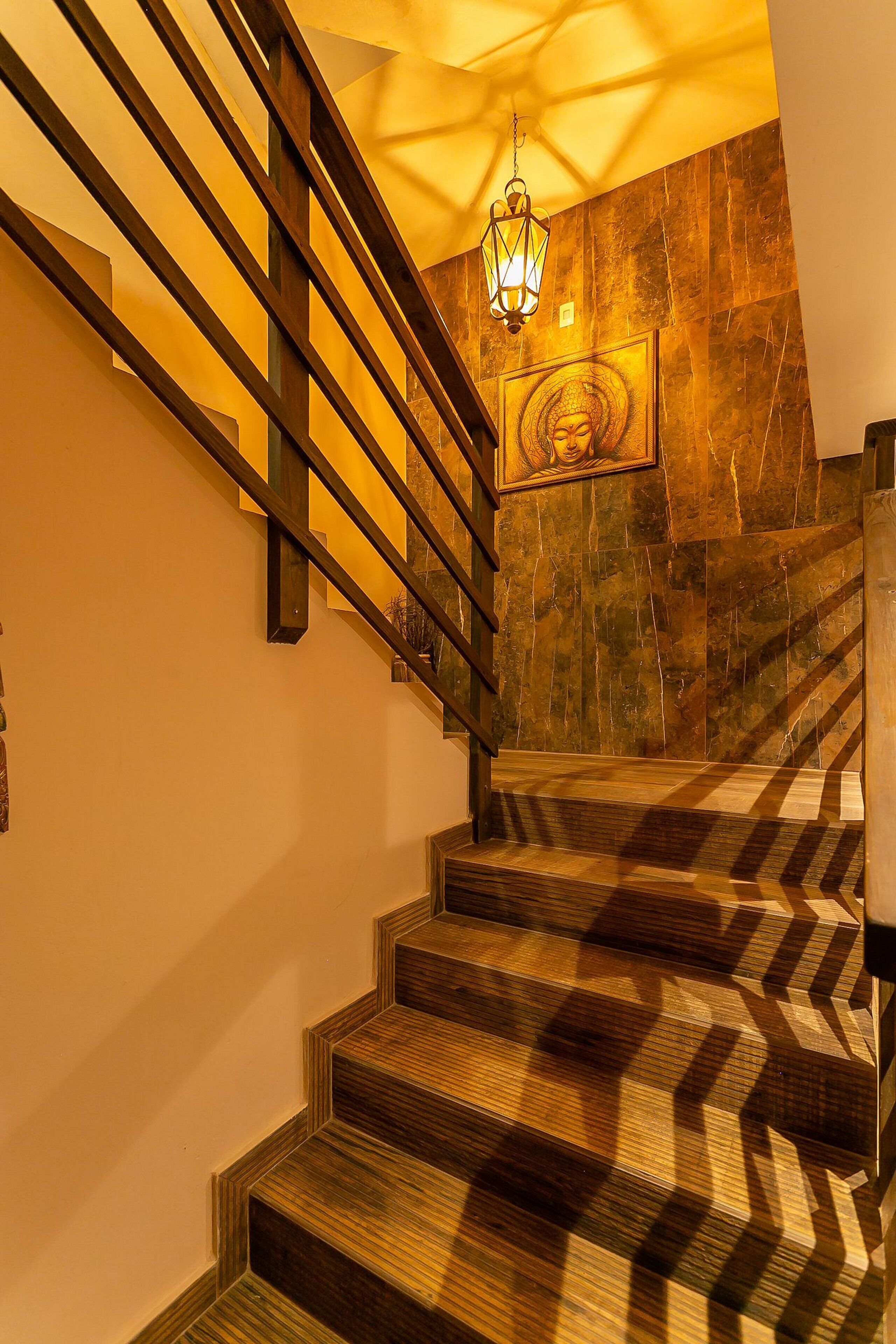Una escalera de madera con barandilla de metal que conduce a una habitación con pared de piedra y una lámpara colgante.
