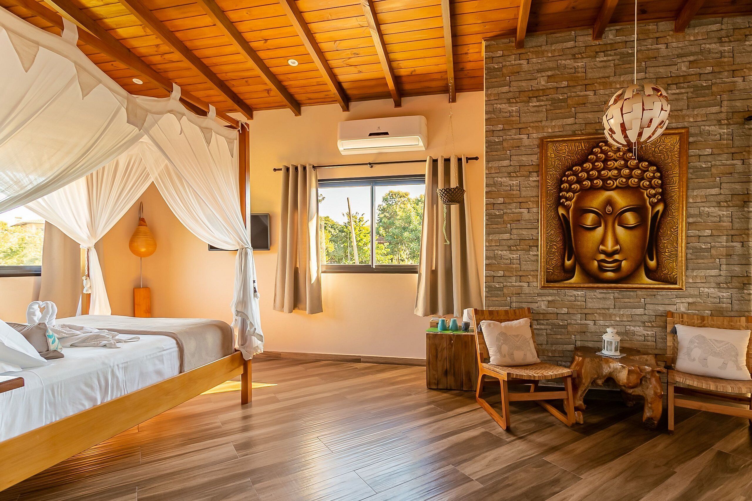 Una habitación con una cama, dos sillas, una estatua de Buda, una ventana y una pared de ladrillo.