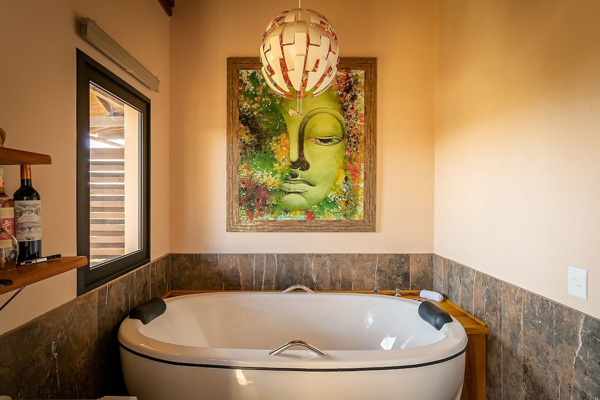 A bathroom with a large bathtub, a wooden shelf with toiletries, a window with blinds, and a wall-mounted artwork.