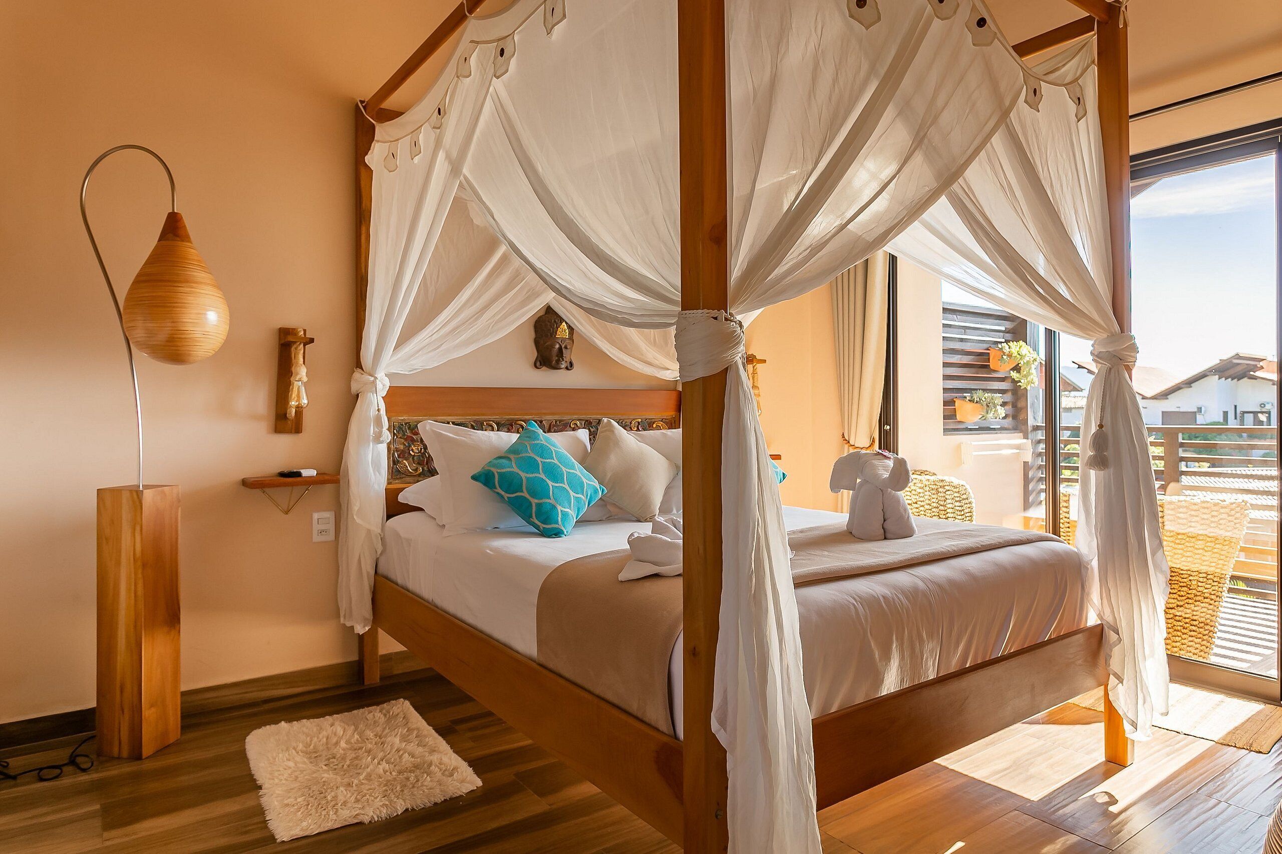 A four-poster canopy bed with white bedding and a blue pillow, a wooden floor, a white rug, and a bedside table with a lamp.