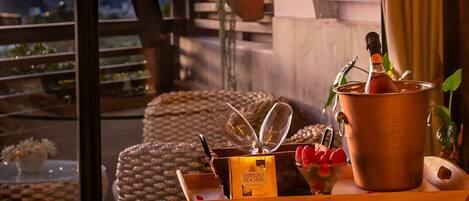 A cozy balcony with wicker furniture, a copper pot with a bottle, and a tray with a Ferrero Rocher chocolate bar and strawberries.