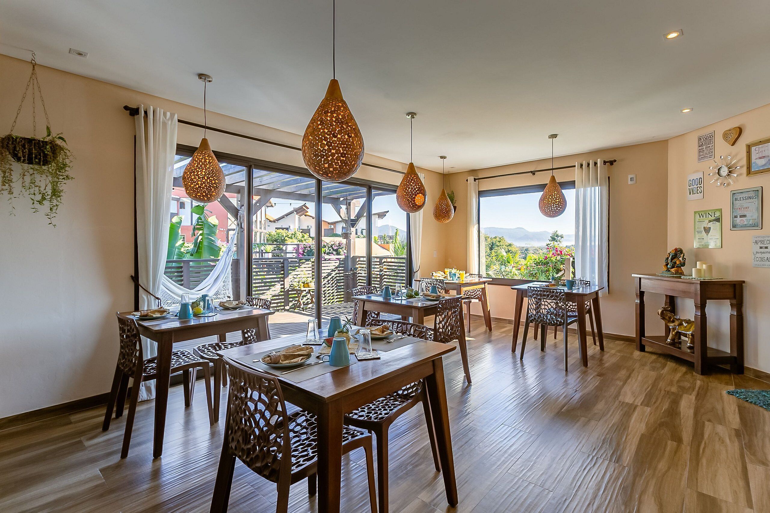 A dining area with wooden tables and chairs, hanging pendant lights, and large windows with curtains.