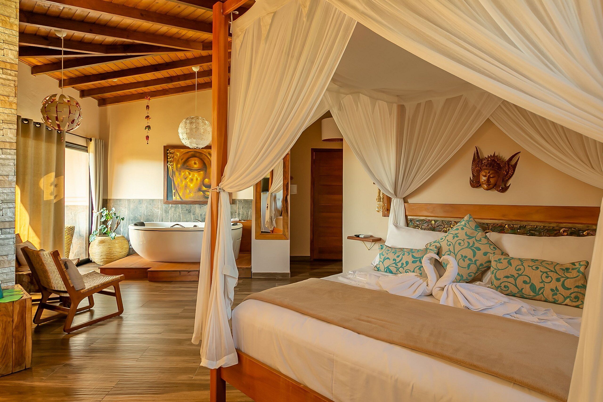 Un dormitorio elegante con cama con dosel, un rincón de estar con una silla, una bañera y una estatua de Buda montada en la pared.