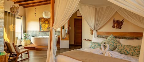 Un dormitorio elegante con cama con dosel, un rincón de estar con una silla, una bañera y una estatua de Buda montada en la pared.