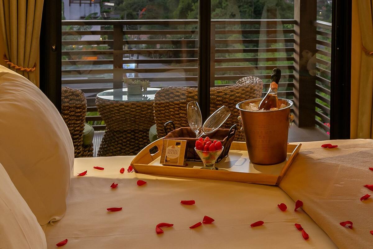 A hotel room with a bed, a balcony with wicker furniture, a table with a bottle of champagne, and a scenic view of mountains and greenery.