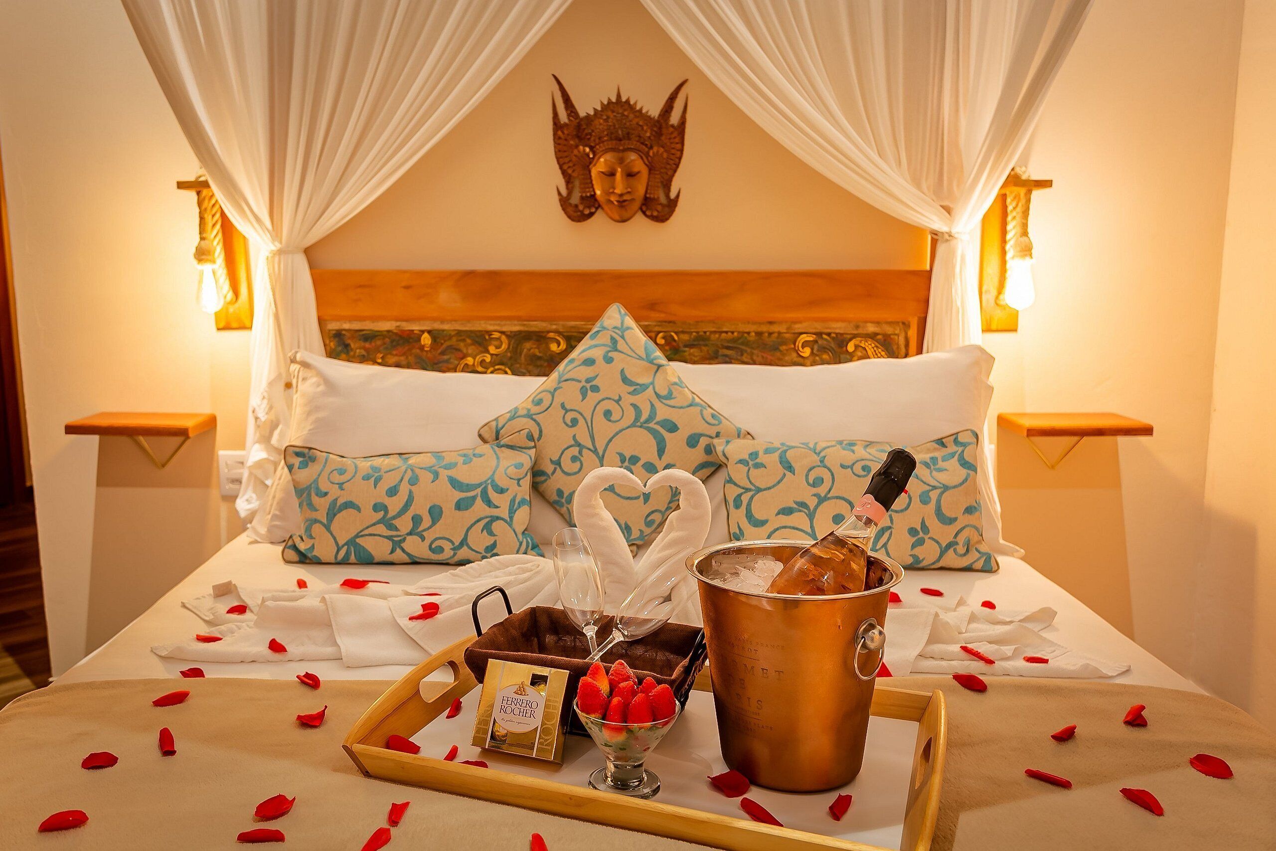 A bed with a canopy, a decorative headpiece on the wall, and a tray with champagne, strawberries, and a bottle of perfume.