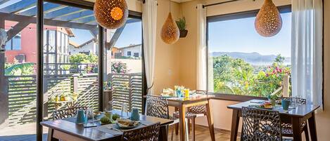 A dining area with a table set for four, two hanging pendant lights, and a view of a garden and buildings outside.