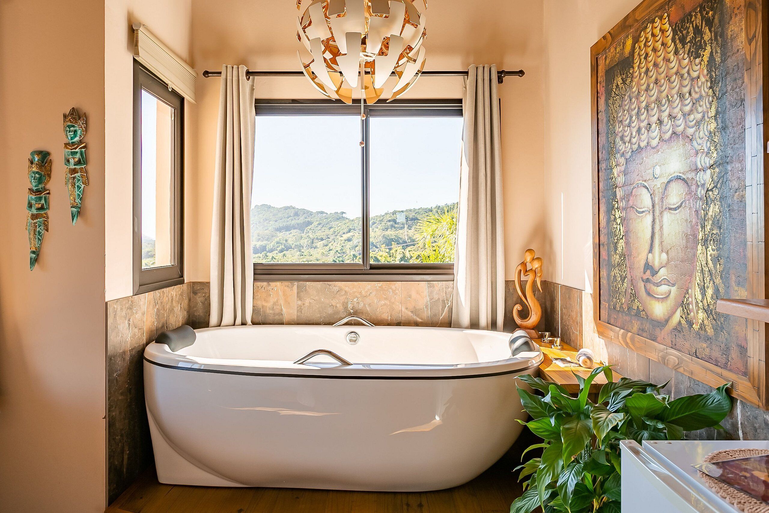 A modern bathroom with a freestanding bathtub, a large window with curtains, a Buddha painting, and a unique chandelier.